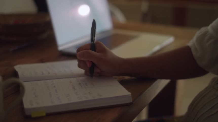 Woman sitting at desk writes in notebook while talking on mobile phone in the room