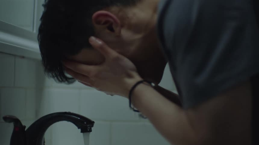 A close up of a young man standing at a bathroom sink looking nervous before splashing water on his face and looking in the mirror