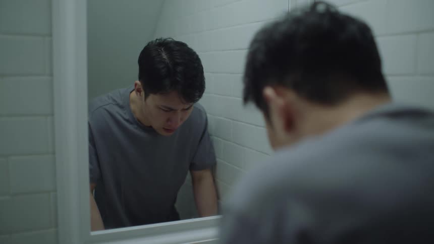 A man appears in front of a mirror and bends down to wash his face then drys it with a small towel looks at himself nervously in the mirror and turns to the door which he opens and walks through to the next room