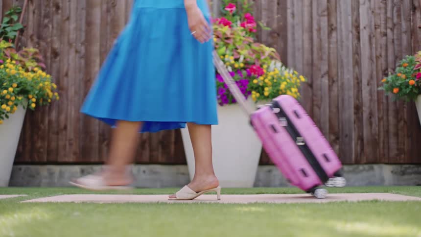 Woman wearing a blue dress carrying a suitcase on a path in front of potted flowers in the garden