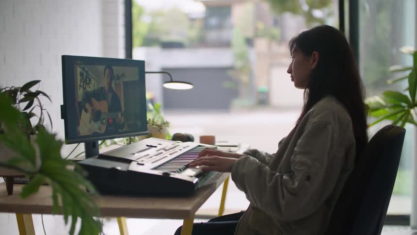 A woman sitting at a desk playing a piano keyboard while on video chat with a man