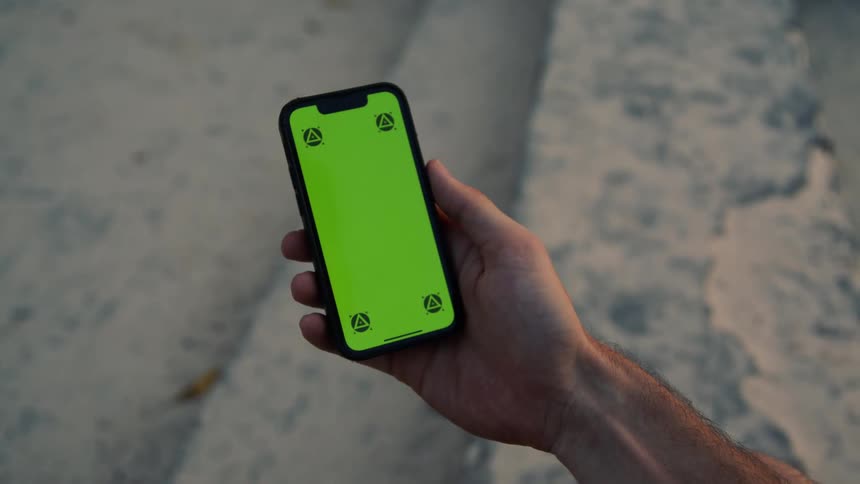 A hand is seen holding a cell phone with a green screen as they scroll with their thumb