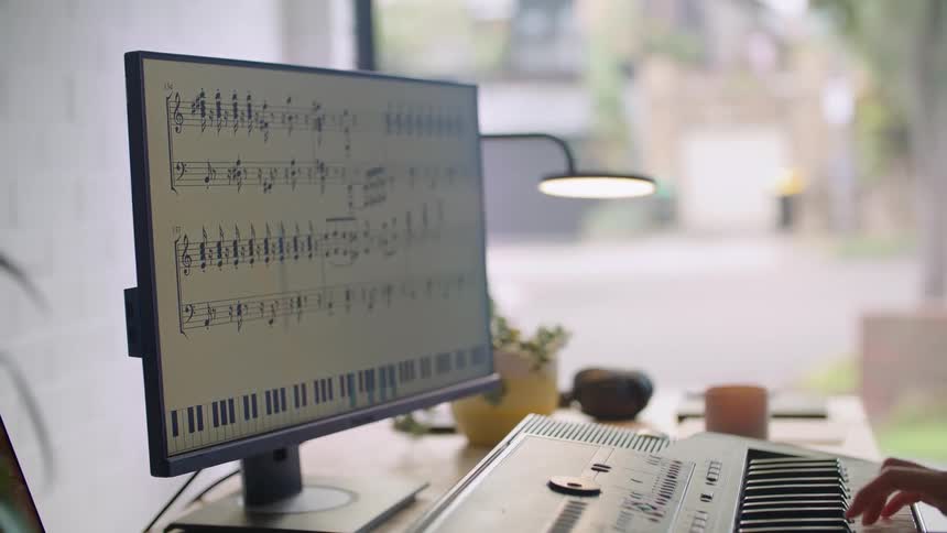 Young woman playing the piano keyboard while sitting at a desk and following musical notes on a pc monitor in the living room