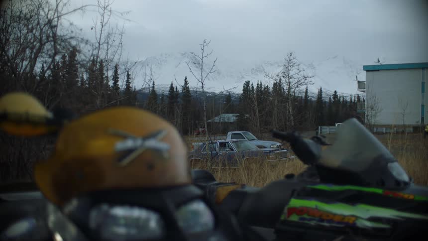 Snowmobiles sitting in a field cars behind them and snow covered mountains in the background