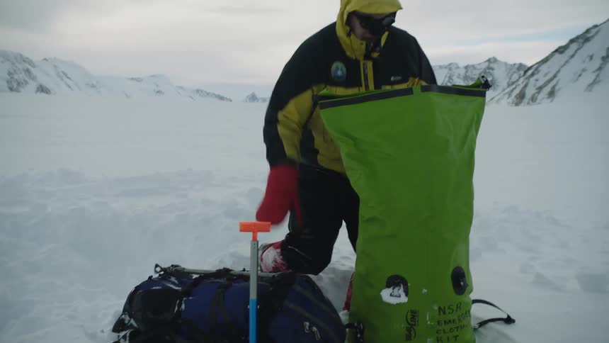 A man in a yellow and black jacket sitting and then standing in snow reaching into his large green bag