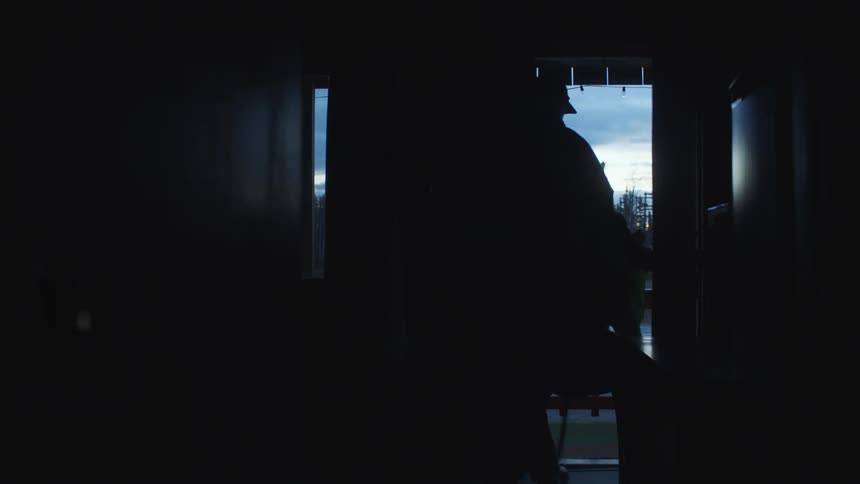 Man wearing heavy backpack and carrying bag is silhouetted in doorway across road from forest