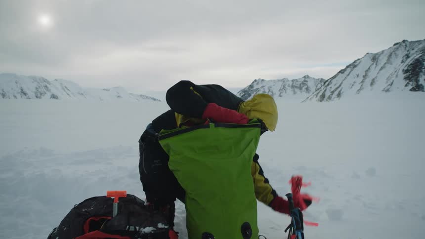 Mountaineer unpacks gear from a green bag over the snow in the mountains at evening