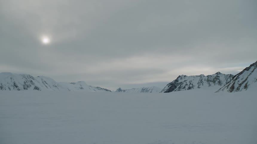 The sun shines over snow covered mountains through a hazy cloudy sky