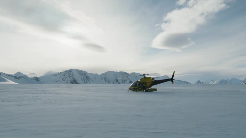 Person stands near helicopter sitting on snow covered plain with mountains in the background