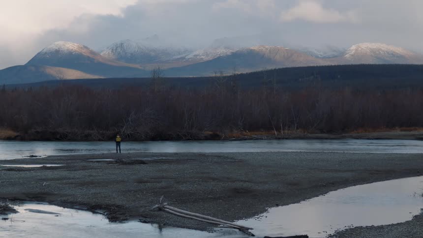 Man wearing a yellow jacket stands on the banks of a river in the wilderness next to the mountains