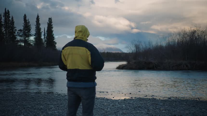 Man with yellow and dark blue coat and jeans watches lake at dusk