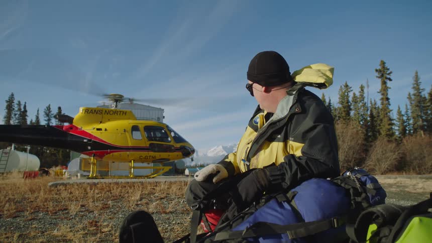 A man knelt down next to bags holding his hat on while he gives thumbs up to the helicopter