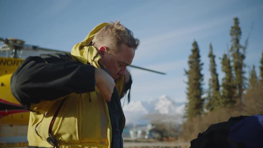 Man standing near a helicopter on a helipad in the mountains puts on a thick yellow jacket