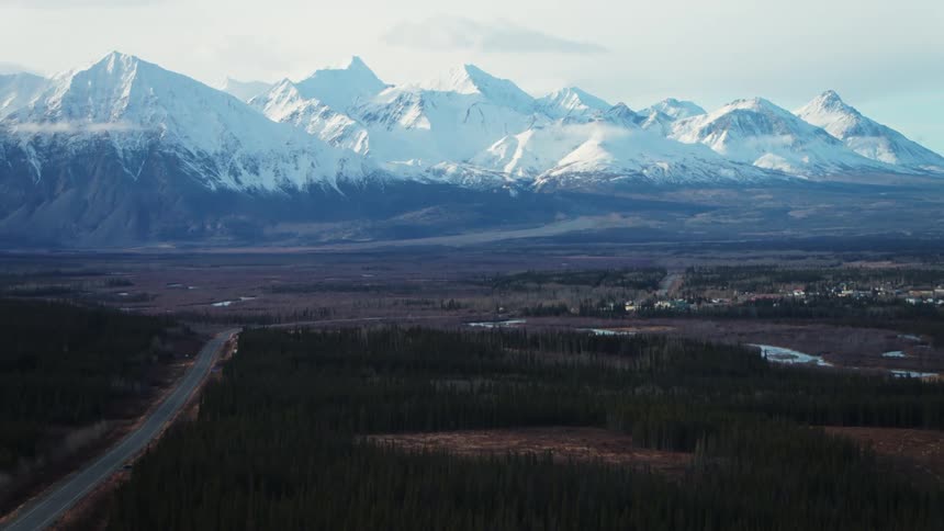An aerial of snow covered mountain tops and a pine forest with a road traveling through it