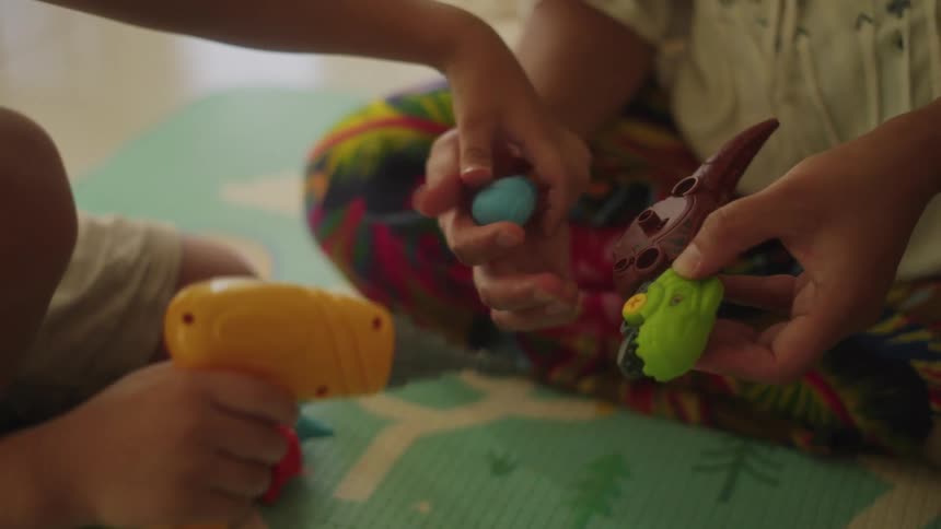 A woman sits on the floor and holds a dinosaur toy while a boy drills it