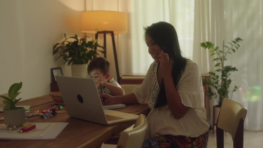 A woman is sitting at a table with a laptop computer writing as she talks on the phone as he little boy is looking at a tablet computer