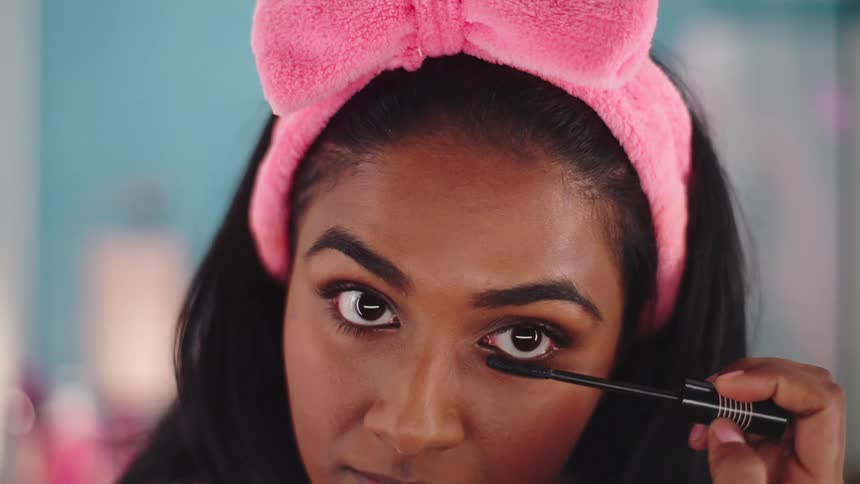 A woman uses a mascara brush to apply makeup to her eyelashes while looking into the camera