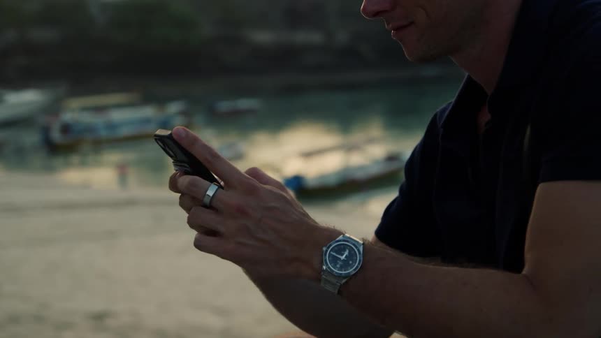 A man who is sitting beside a lake in the evening smiles as he looks down at his phone and begins rapidly texting on it