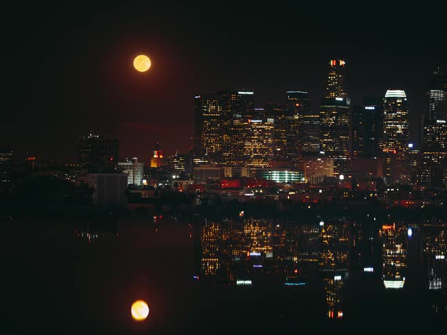 A time lapse of city buildings at night as a full moon rises up and over the buildings