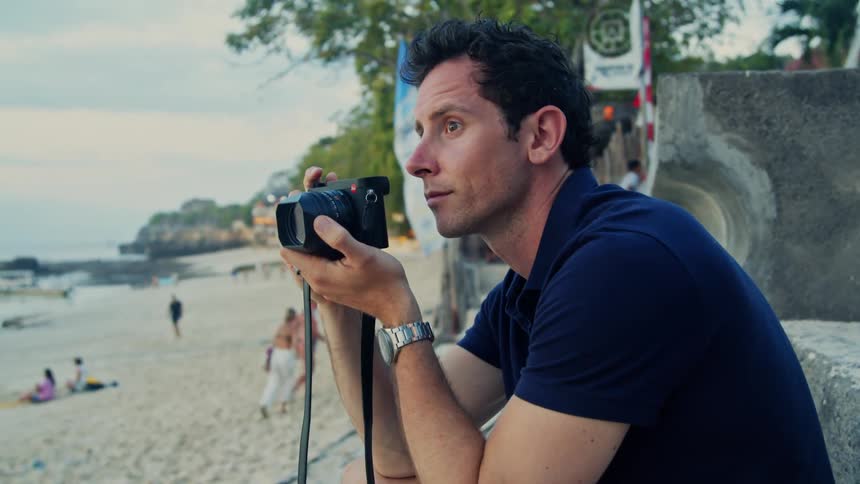 A man raises a camera to his face to take a photograph while sitting on a beach during the day