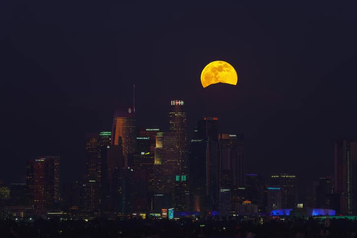 The sun sets and the moon rises over the skyscrapers of a city
