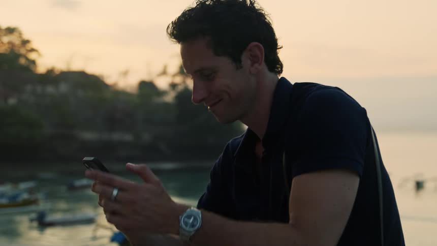 A man wearing a black tshirt types on his phone while sitting near the water outdoors