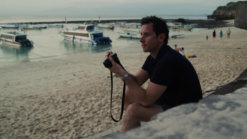 Male photographer sitting near beach takes pictures with moored boats and beachgoers in the background