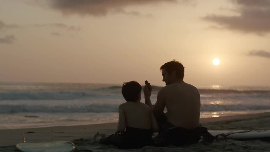 Father hands son gift as they sit on beach between surfboards under a red tinted sky