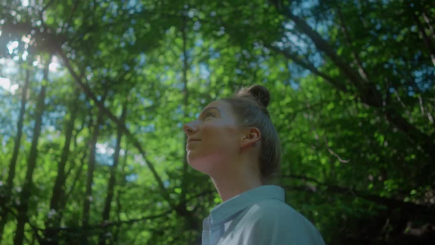 A teenage girl is standing in the woods looking at the trees
