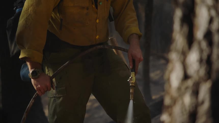 Firefighter wearing yellow protective gear hoses the ground on the forest