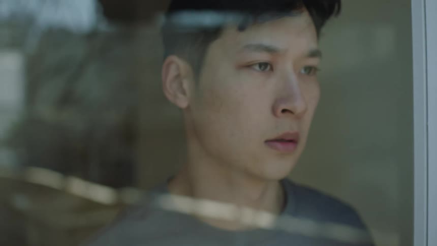 A teenage male stands in a window and sadly peers outside before turning to leave