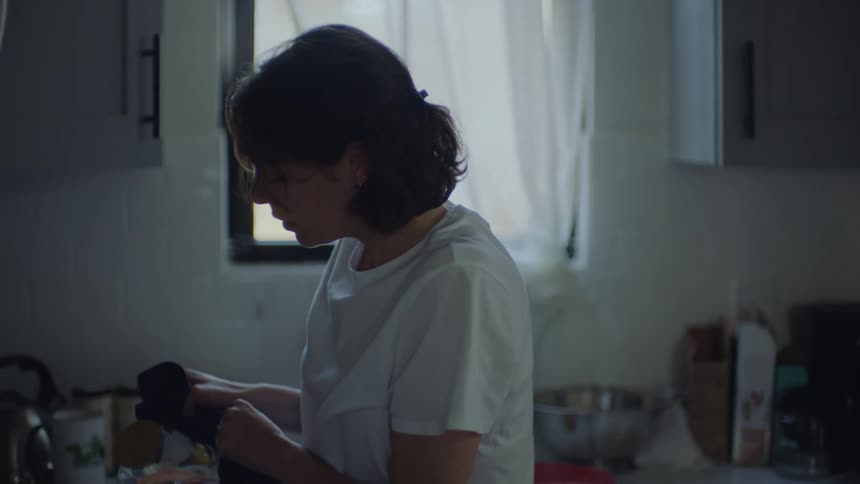 Woman leans against kitchen counter before standing to pour hot water then leaves with beverage