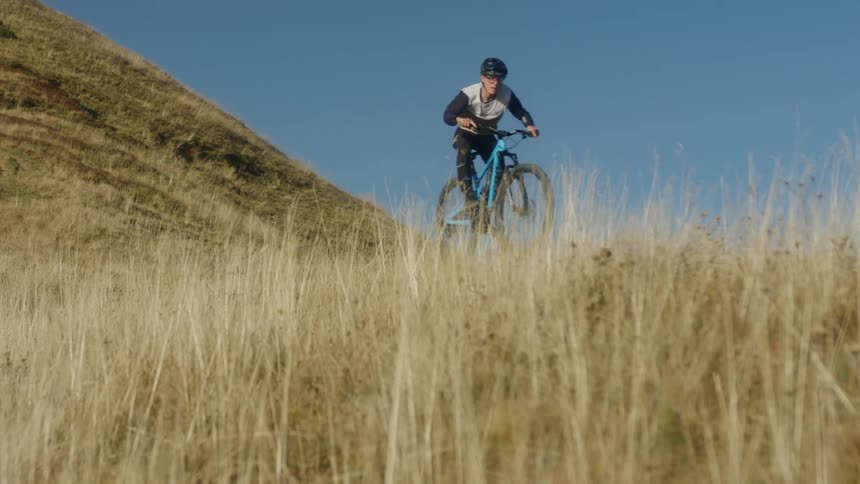 A males rides a bike through tall grass near a hill