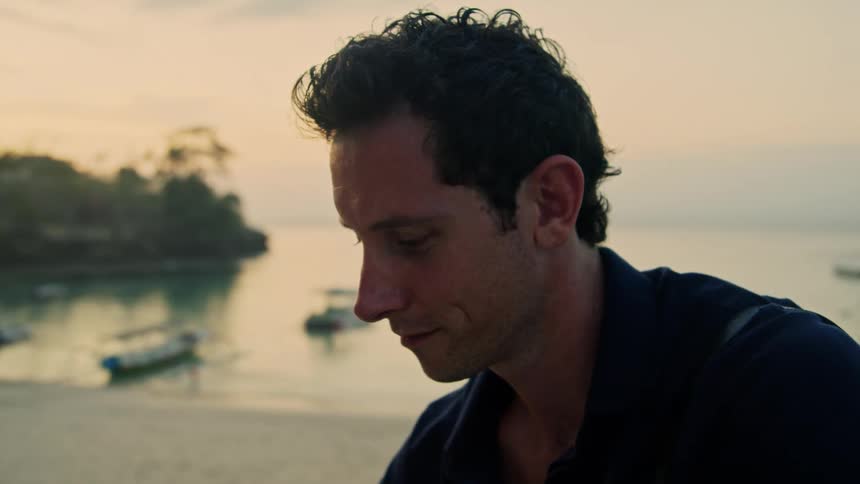 A man is texting and sitting on the beach at dusk