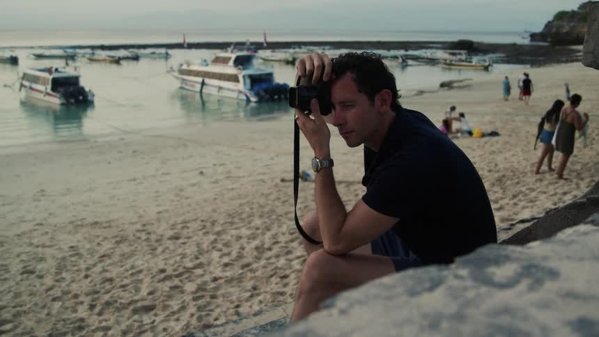 Man in black shirt and shorts takes photo with camera on the beach next to sea with boats