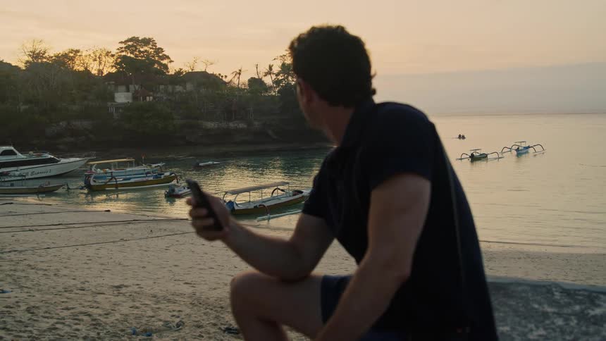 Man in black shirt and shorts uses smartphone on the beach at sunset