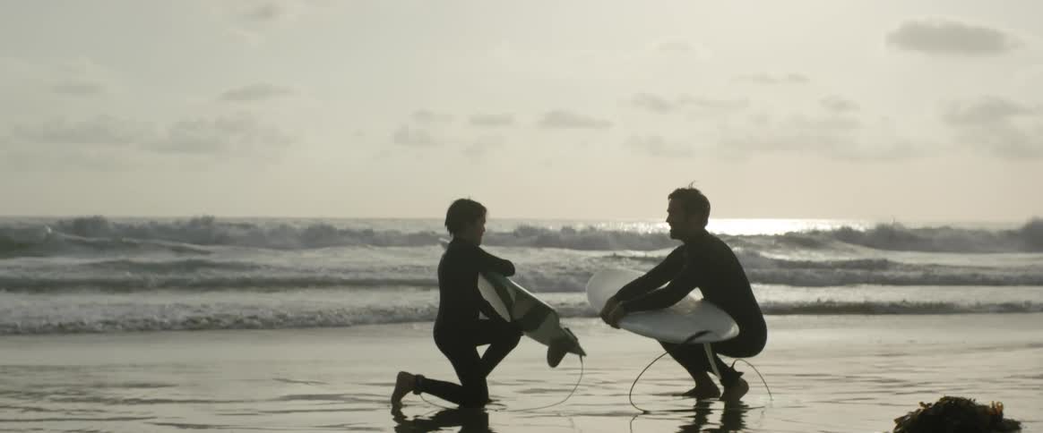 Father and son are kneeling on a beach with surfboards and talking