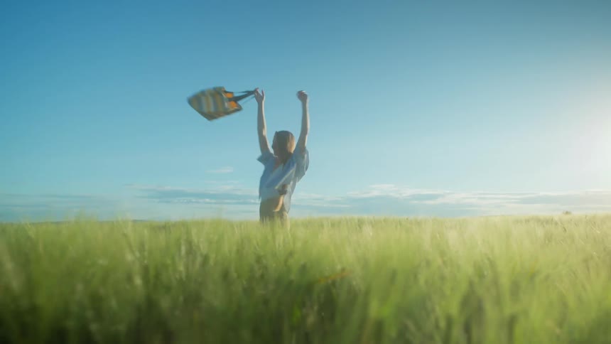 Happy woman holding a bag with arms outstretched spinning around in a grassy field
