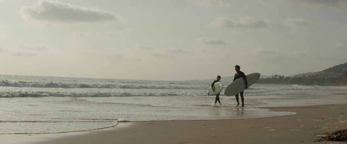 Father and son carrying surf boards walking down the beach through the water