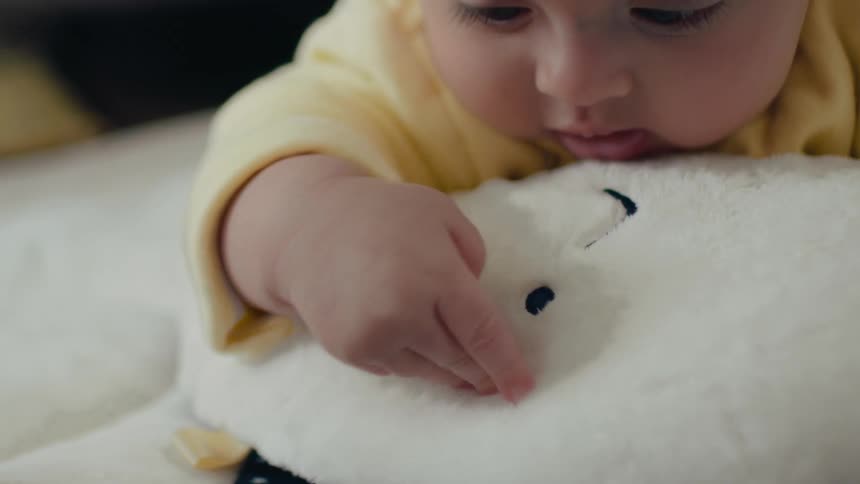 Baby wearing yellow leans on flat stuffed toy while lying on bed