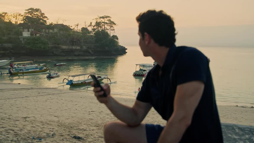 A man is sitting on the beach as he uses his cell phone and there are small boats in the water along the shore
