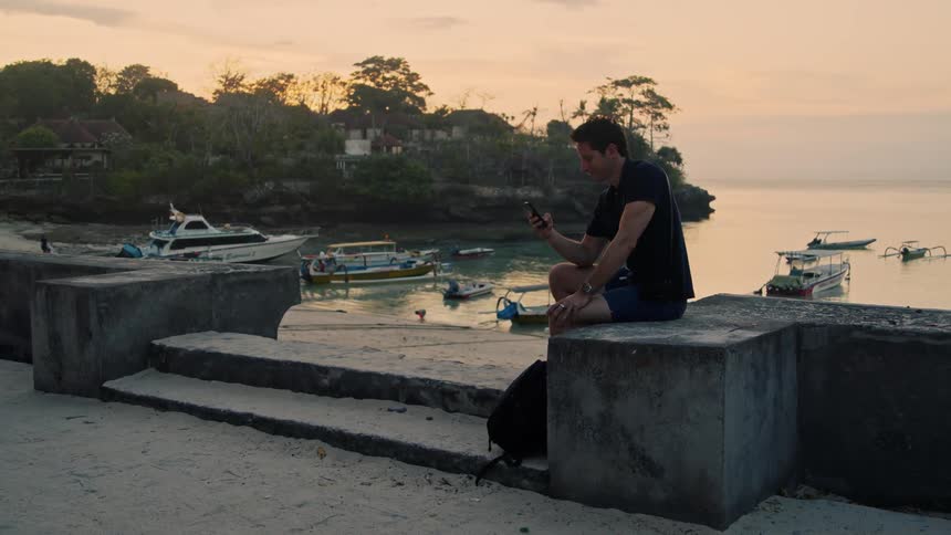 Young man sitting near a pier in the tropics texting on his phone at sunset