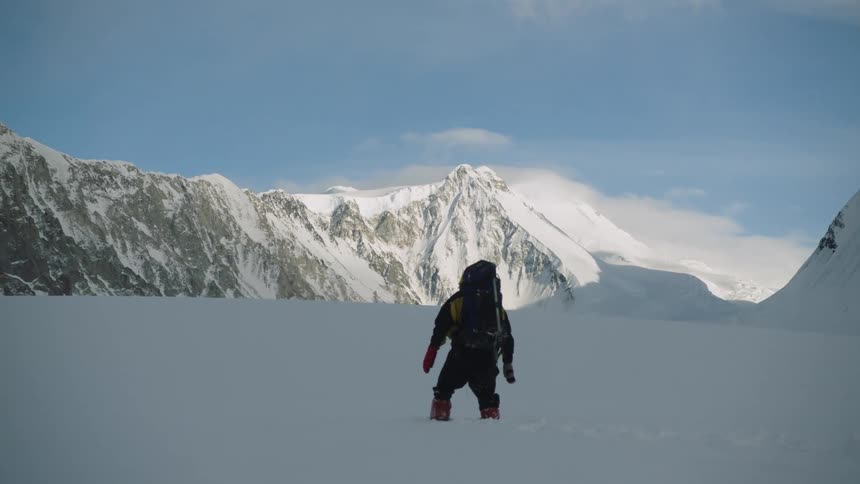 A person dressed for extreme weather struggles to cross a field of deep snow in the mountains during the day