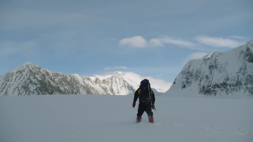 A man bundled up in a hooded winter coat and boots wearing a large backpack is carefully gets his footing as he makes his way through deep snow with mountains in the distance