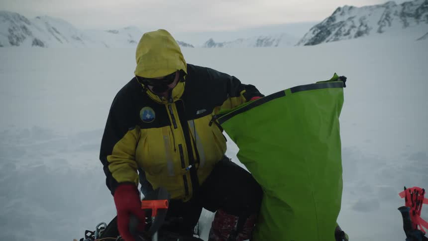 Man wearing cold weather gear packs harness and other items into large sack while half kneeling in snow with mountains in the background