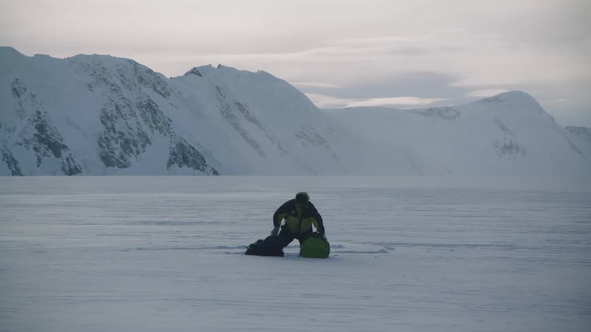 A hiker tries to condense two backpacks while kneeling in the snow with mountains in the background