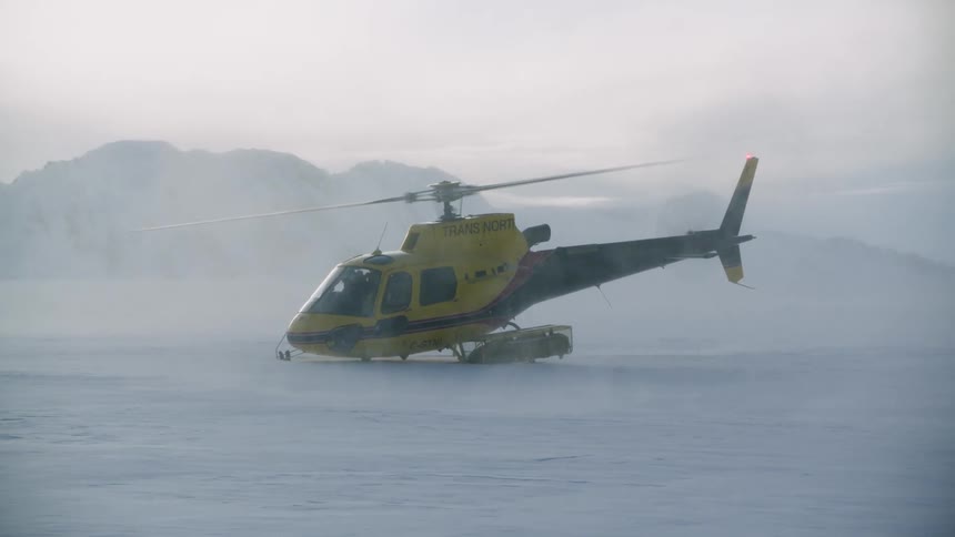 A helicopter is sitting on snow covered ground and snow flies in the air as the rotor spins and there are mountains in the distance