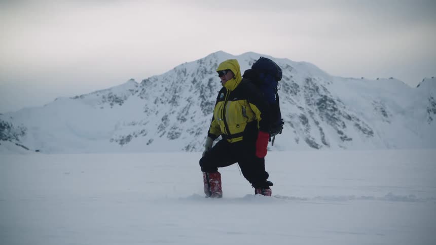 A man bundled up in a hooded winter coat and boots wearing a large backpack is carefully gets his footing as he makes his way through deep snow with mountains in the distance