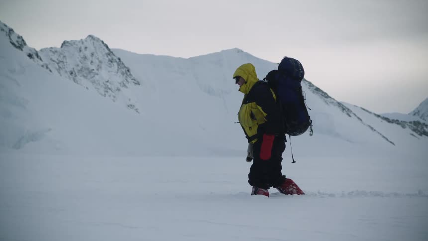 A man bundled up in a hooded winter coat and boots wearing a large backpack is carefully gets his footing as he makes his way through deep snow with mountains in the distance