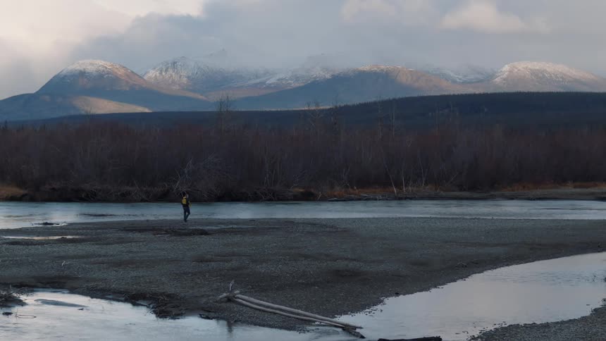 Man walks along sand bar across water from leafless woods backed by mountains